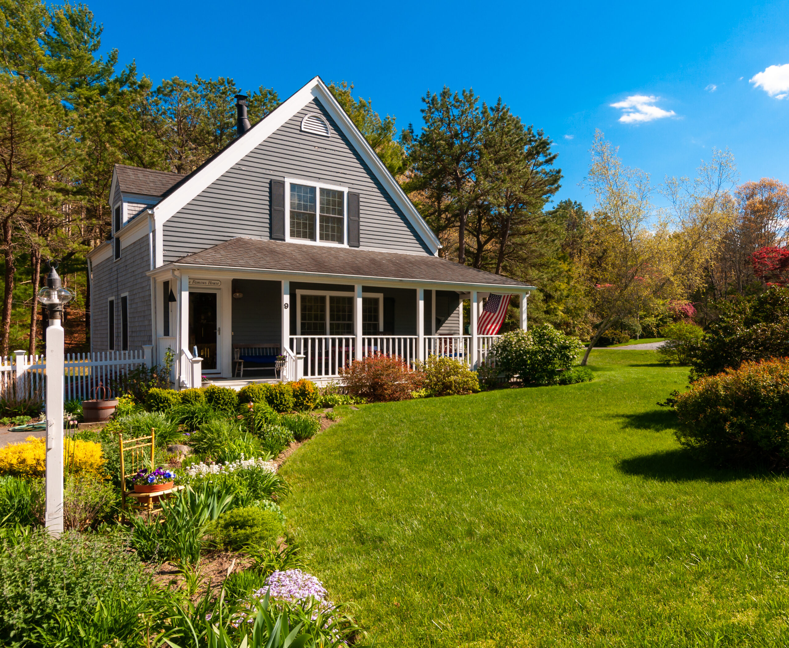 An American flag flies from the covered front porch of a small single family home with well maintained grounds and gardens on Cape Cod in Massachusetts.